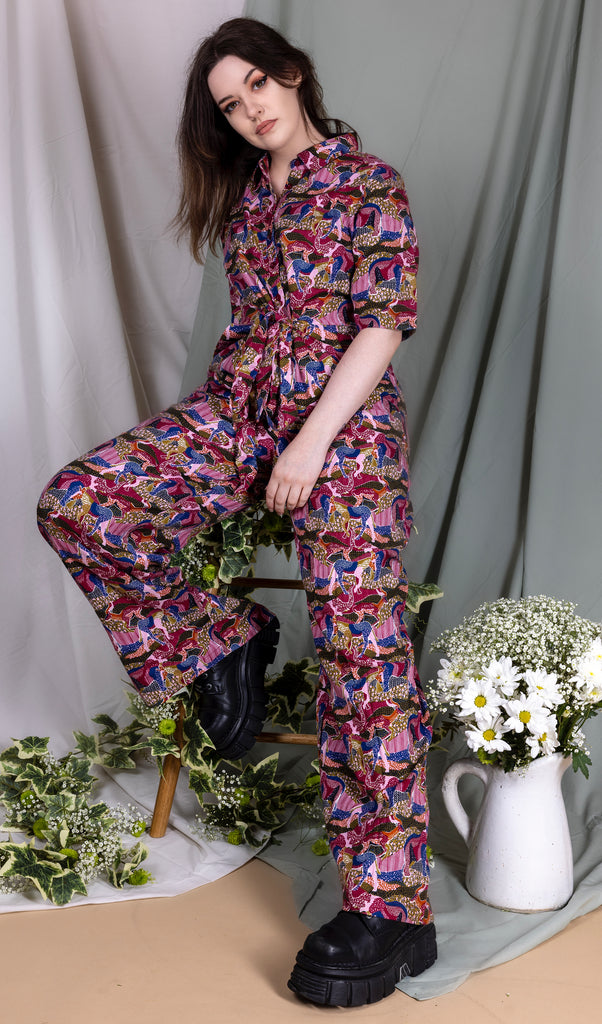 Alice is wearing the autumn safari jumpsuit sat leaning back on a stool in front of a draped sheet backdrop with various flowers and vines on the floor and stool. She has one leg propped up on the stool and one leg out towards the camera to highlight the jumpsuit leg. The print features blue, brown, orange, pink, red and white patterned safari animals all over. The jumpsuit buttons down with a collar and belt tie at the waist.
