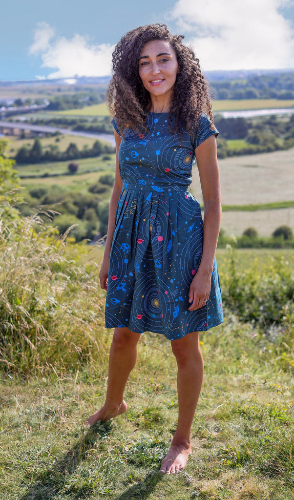 Rochelle is stood on a grassy hilly area wearing the retro rockets solar intergalactic tea party dress. They are facing towards the camera smiling.