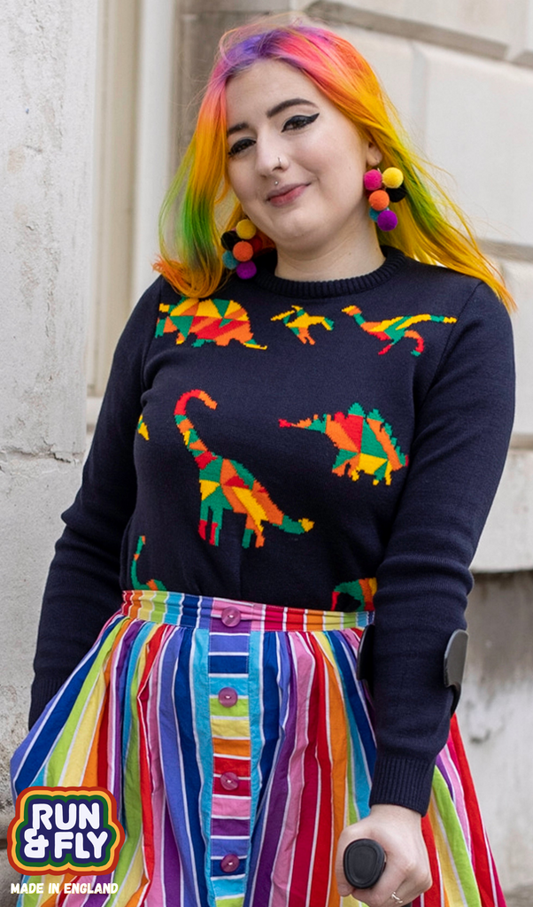 Eliza is stood in front of a white brick wall wearing the navy rainbow dinosaur jumper with a rainbow skirt and rainbow earrings to match their rainbow hair. They are posing facing the camera with their hand holding their walking aid in front of them whilst the other is in their skirt pocket. Photo is cropped from the hips up.