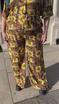 Video of Luisa Christie wearing the midnight blues sunflower jumpsuit, floral pattern in yellow, green, orange and deep blue. Model is smiling and posing around in a sunny Brighton street, wearing sunglasses and black boots with arms by sides.