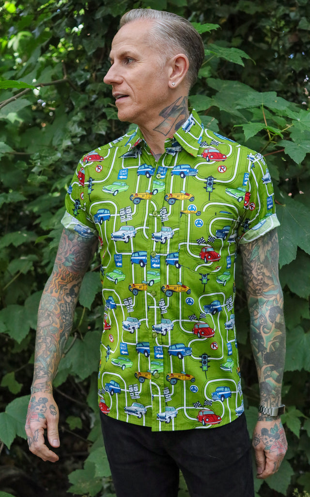 Jim is stood outside in front a bushy area wearing the kiwi car highway short sleeve shirt with black jeans. They are heavily tattooed and posing facing the camera but looking off to the distant left. Photo is cropped from the thighs up.