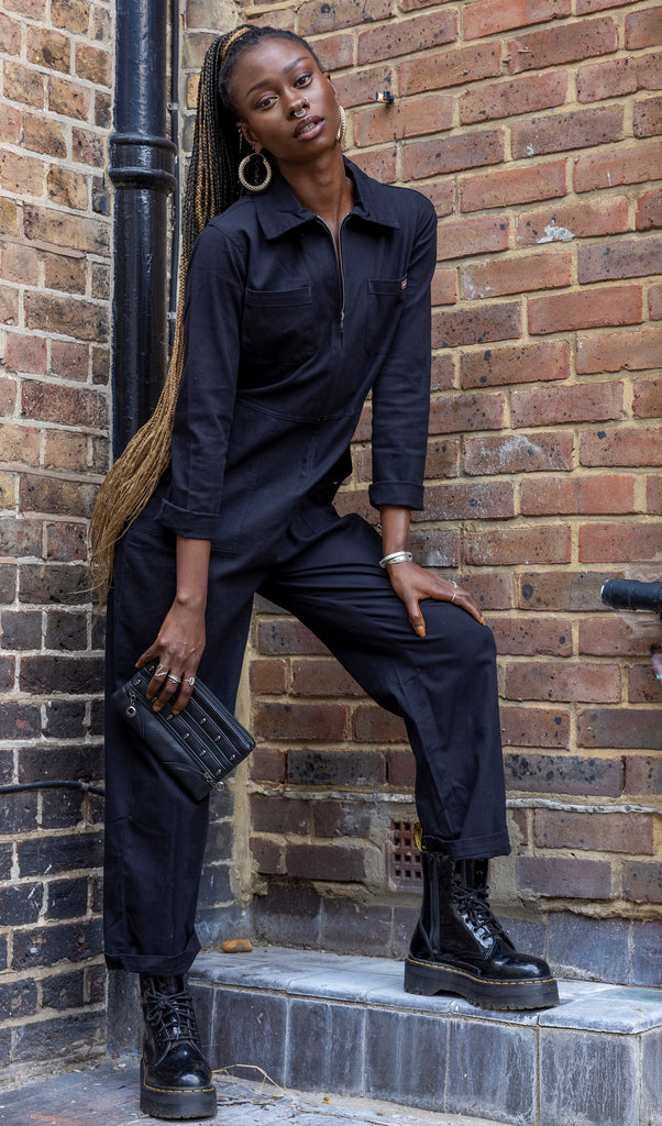 Yolande is stood outside in front of a brick wall corner wearing the black stretch twill boiler suit with black boots and holding a black clutch bag from gothx. She is facing forward, has one leg up on the step and leaning forward on that leg whilst looking to camera. The boiler suit is all black with a silver front zip, collared top, two top front pockets, side pockets and room to roll if needed. 