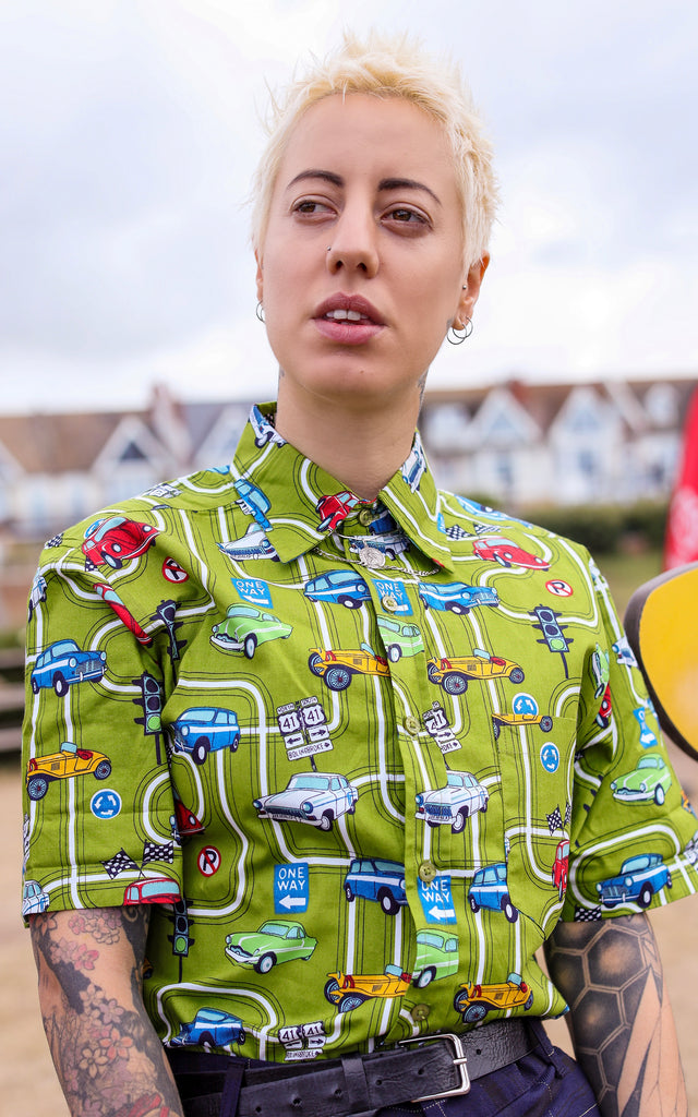 Yo is stood near a beach wearing the kiwi car highway short sleeve shirt with blue tartan trousers. They are facing the camera and looking off to the left. Photo is cropped from the waist up.