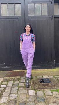 white femme model with black hair and tattoos is stood outside in Hove in front of a black door wearing Lavender Stretch Corduroy Dungarees paired with Cat Chorus Short Sleeve Tee and silver boots. Model is posing looking with hands in the dungaree pockets, walking toward the camera and showing the stretch in the bib of the dungarees.