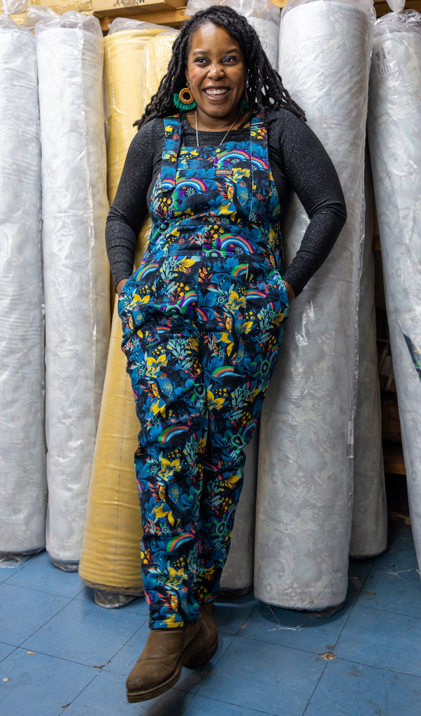 Ama is standing in a warehouse next to rolls of fabric, smiling with hands in pockets wearing the black jurassic stretch twill dungarees with a black long sleeve top underneath and brown boots. 