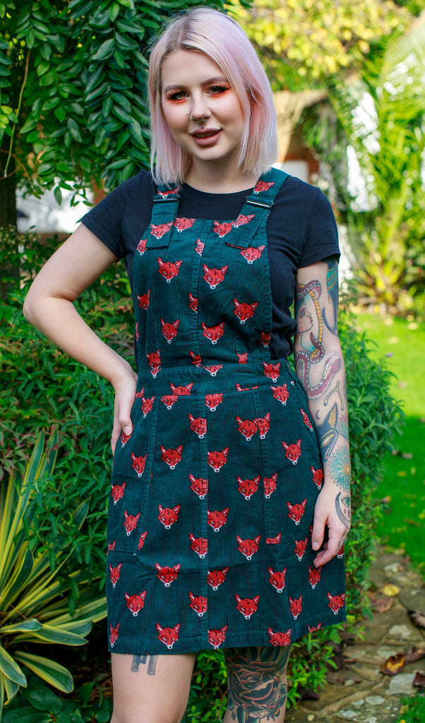 Florence is wearing the fox stretch corduroy pinafore dress, featuring red fox heads on forest green, paired with a black short sleeved tshirt. Tattooed model is smiling and looking at the camera, in a green garden with a hand on her hip.