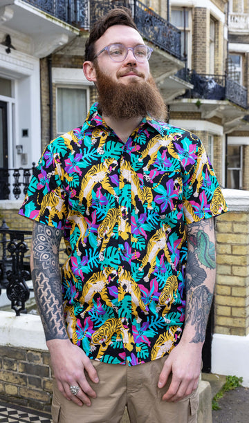 Tattooed model is wearing Black Tiger Lily Stretch Short Sleeve Shirt, paired with beige trousers. The collared shirt has a black background colour with all over orange tiger print and various foliage in pink, purple and green. Model is smiling and looking into the distance.