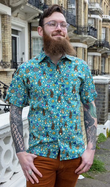 Dec is wearing the Bunny Meadow short sleeve blue shirt with floral pattern, worn with brown corduroy stretch flare trousers. Model is looking away from the camera outside of a street.