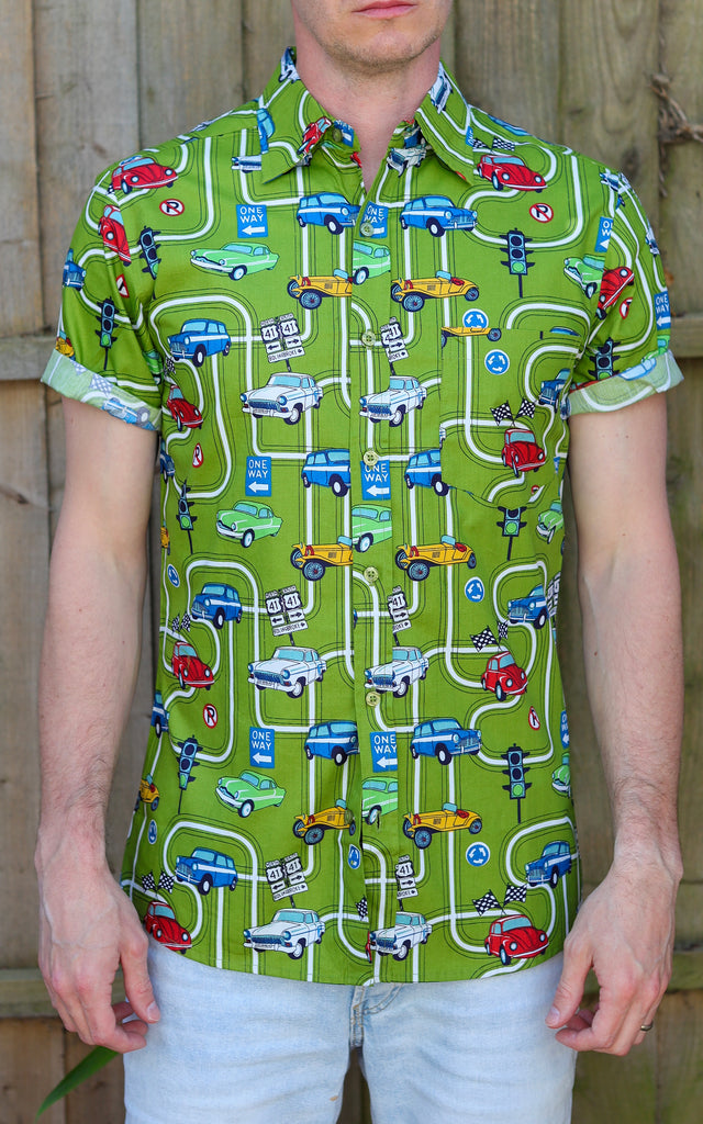 Model is stood in front of a wood fence wearing the kiwi car highway short sleeve shirt with light denim jeans. They are facing the camera and photo is cropped from the thighs up.