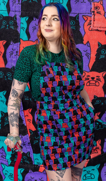 Eliza is wearing the cat chorus stretch twill pinafore dress with a dark green leopard print tshirt underneath. They are standing leaning on their red walking aid with one hand in the dress pocket whilst smiling looking left. They have rainbow mid length hair, eyeliner and tattoos with a red walking aid and iridescent handle. The cat chorus print is an all over repeating pattern of sky blue, peachy red, purple and black cats pulling various faces and a sporadic sky blue bird.