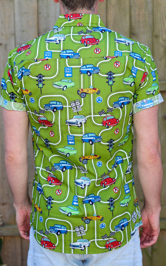 Model is stood in front of a wood fence wearing the kiwi car highway short sleeve shirt with light denim jeans. They are facing away from the camera and photo is cropped from the thighs up.