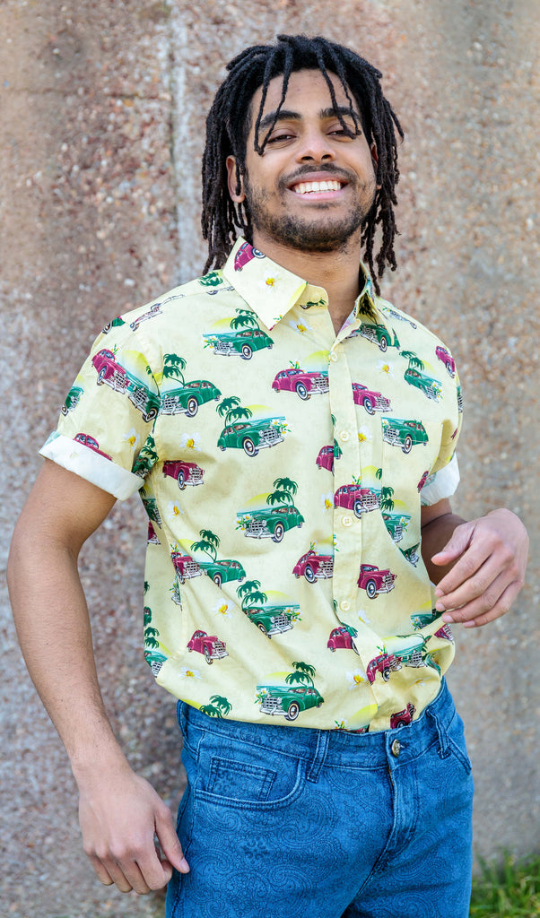 Model is stood outside in front of a concrete wall wearing the Cuba short sleeve shirt with denim paisley flares. They are facing the camera with one hand up by their front with the other resting by their side, they are looking into the camera and smiling. Photo is cropped from the thighs up.