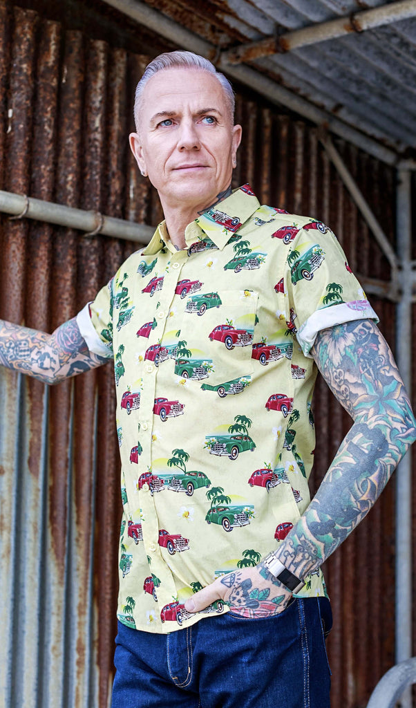 A heavily tattooed model is stood in front of a rusted metal wall wearing the Cuba short sleeve shirt with denim jeans. They are facing the camera with one hand resting up on the metal wall and the other in their front pocket. They are looking off to the right. Photo is cropped from the hips up.