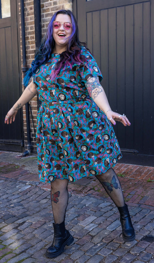 Luisa Christie is wearing the Hedgehog tea dress featuring cute hedgehog print on blue with pink, paired with black fishnets and chunky boots. She is smiling and posing in a cobbled street in front of black doors. 