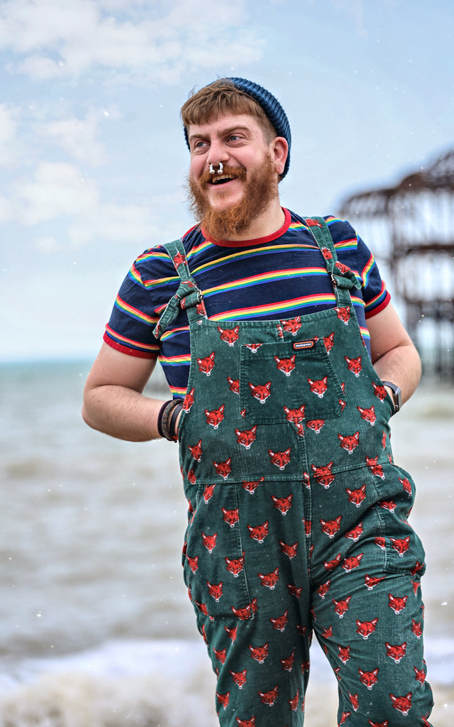 Model standing by the sea wearing a beanie, green fox dungarees and the Retro Rainbow Block Striped T Shirt
