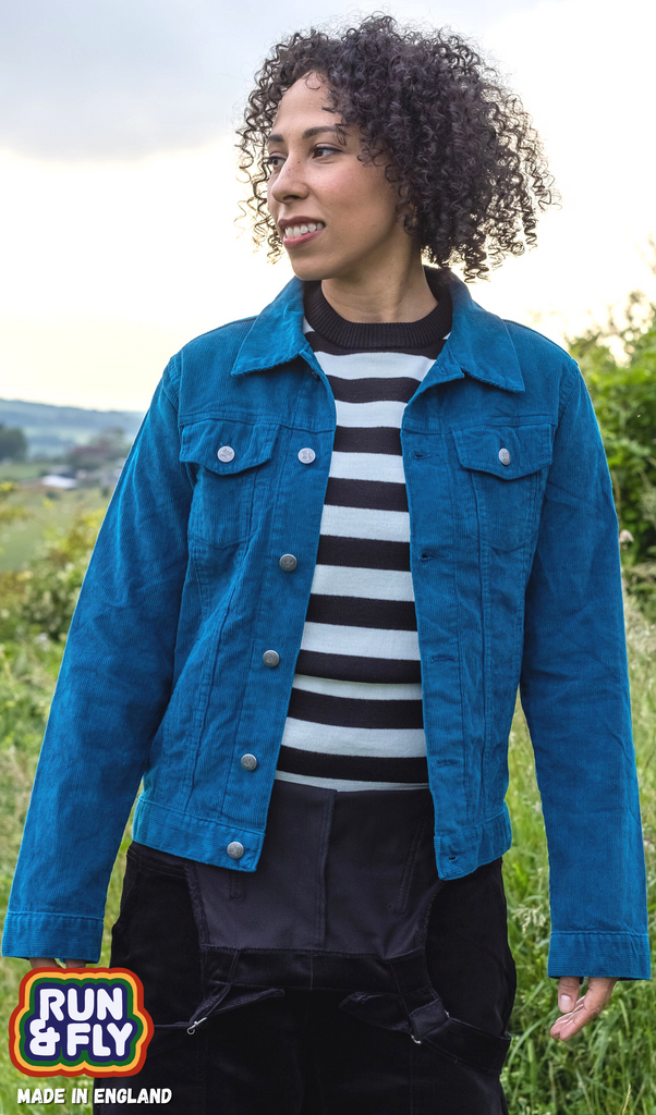 Amy is stood in a field area wearing the retro vintage ink cord unisex western jacket with a striped tshirt and black dungarees. They are facing forward with their arms out by their sides whilst smiling to the left. Photo is cropped from the hips up.