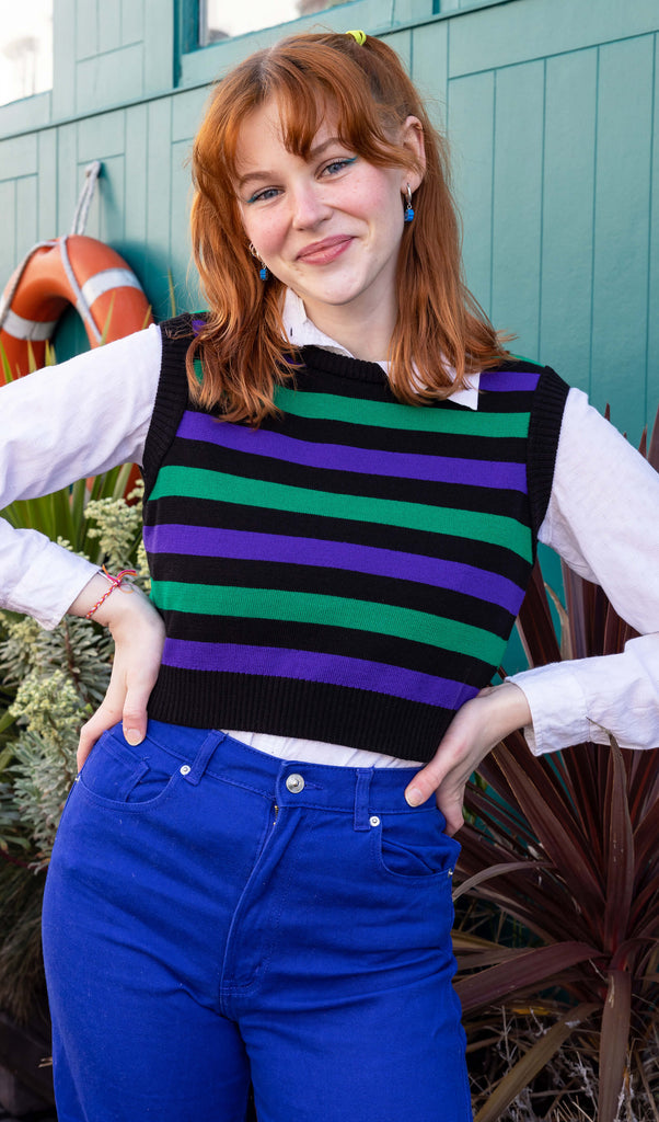 Tasha a tall red haired femme model is wearing a white shirt and striped tank with blue jeans against a mews background