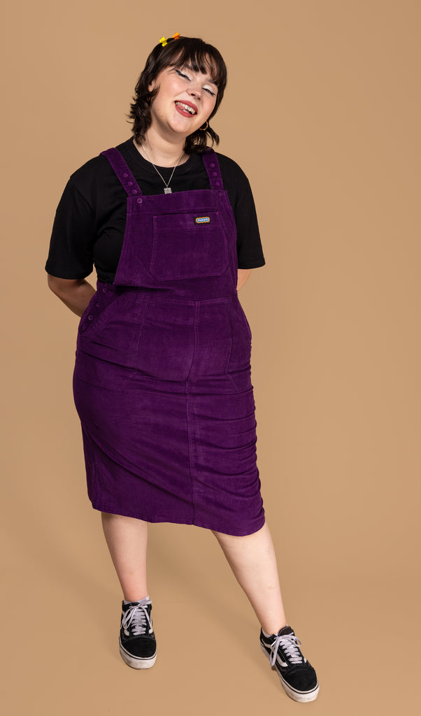 Rebecca is stood in front of a beige background wearing the purple stretch corduroy long pinafore dress with a short sleeve black tshirt underneath and black trainers. She is leaning back on one leg with both hands behind her waist whilst smiling to camera. The long pinafore is all purple with button down straps.