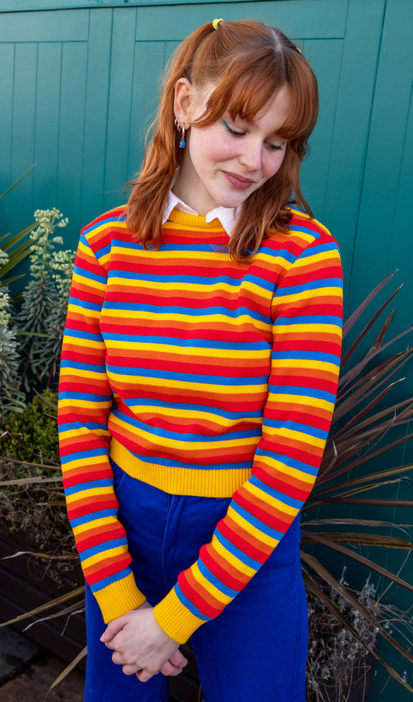 Tasha is wearing the Rainbow Primary Colours Striped Cropped Jumper with a white collared shirt underneath and blue jeans. She is stood outside in front of a blue wall with plants and is posing with their hands together and looking down facing the camera.
