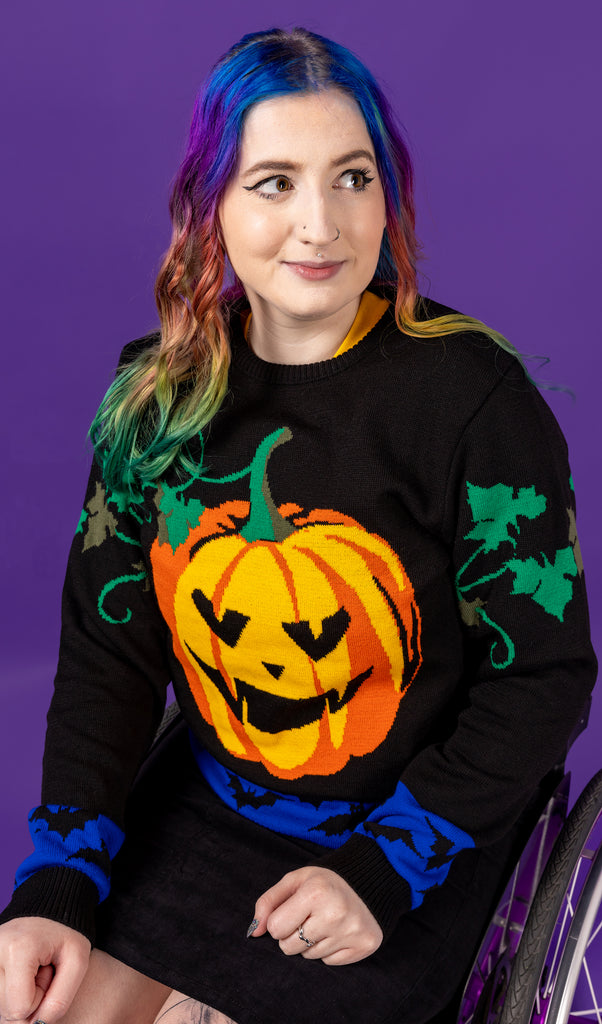 Eliza is sat in their wheelchair wearing the Halloween: Pumpkin Jumper with a black pinafore underneath in front of a purple background. They are facing slightly left looking off to the right smiling with both hands resting in their lap. The pumpkin jumper is all black with a carved pumpkin in the centre, green vine leaves around the top of the arms and a blue bat stripe along the bottom hem and arm cuffs.