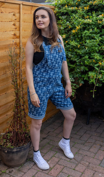 Amy is stood in a garden wearing the blue daisy printed denim stretch dungaree shorts with a black vest top and white trainers. She is facing the camera and looking off to the left.
