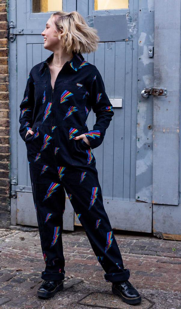 Amy is standing in front of grey warehouse doors wearing the black lightning rainbow stretch twill boiler suit with front zip, pockets and collared neck. Model is looking away from the camera with hands in pockets and smiling