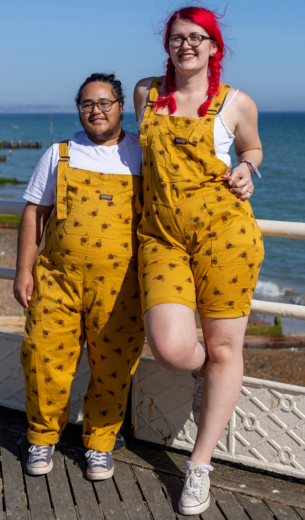 A couple stood on a pier in front of a blue sea are wearing the matching bee stretch twill dungaree shorts and dungarees. One has mid length red hair and both are smiling wearing glasses.