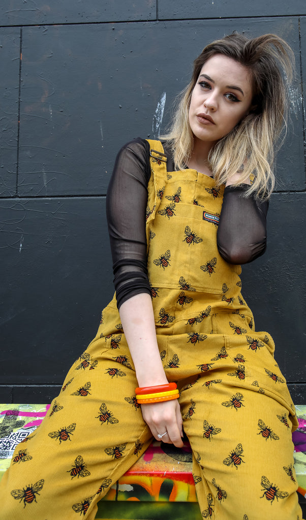 Alice is sat outside in front of a black brick wall wearing the gold corduroy dungabee dungarees with a long sleeve black mesh tshirt underneath and multicoloured bracelets. They are facing the camera and posing with one hand behind their head and the other resting in their lap. Photo has been cropped from the knees up.