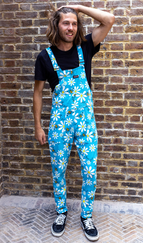 Rudi is wearing the bee free blue stretch twill dungarees, featuring white daisies and yellow bees on blue, paired with a black shot sleeved t-shirt and black trainers. Model is posing with one hand by sides and one hand through long hair, in front of brick wall. 