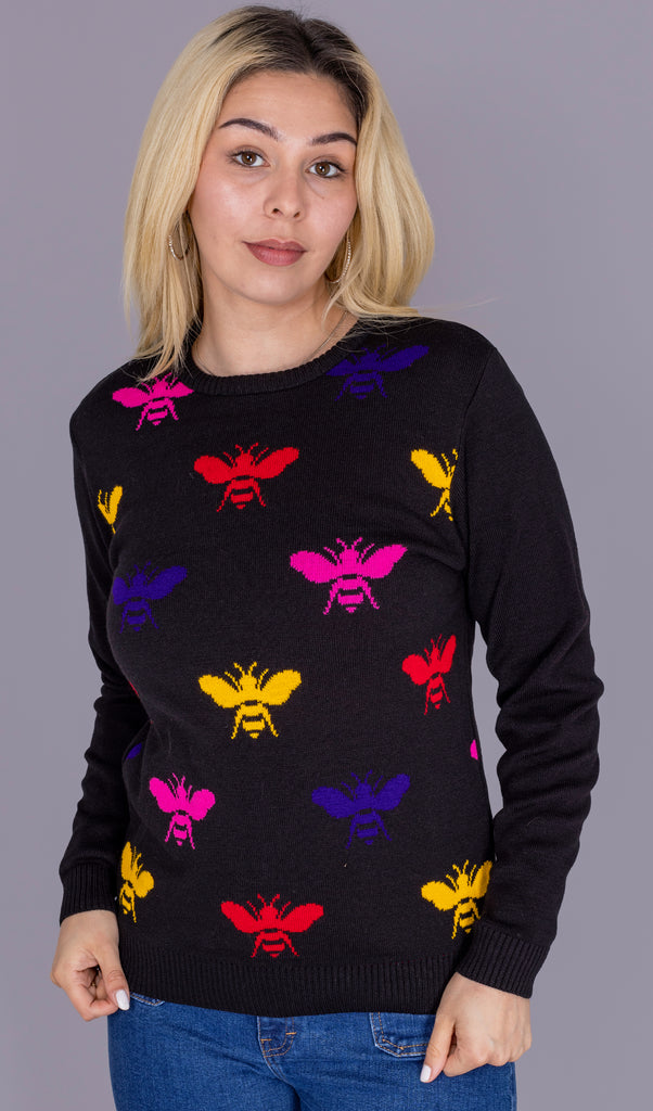 Shannon is stood in front of a grey studio background wearing the rainbow bee jumper with blue flares. They are facing forward posing with both hands holding onto the bottom of the jumper whilst smiling to camera. Photo is cropped from the hips up.