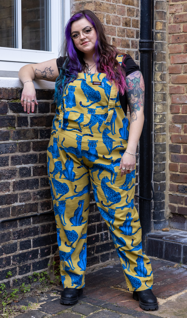 Luisa is standing in front of a brick wall wearing the big cats stretch twill dungarees in yellow and blue with front pockets, paired with a black tshirt and chunky boots. Model is facing the camera and smiling while leaning against the window ledge.