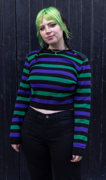 Model with green and black hair wearing Black Striped Purple and Green Cropped Jumper paired with black jeans. The jumper is a cropped style and has a black background colour with chunky muted green and purple horizontal stripes across it. Model is smiling at camera.