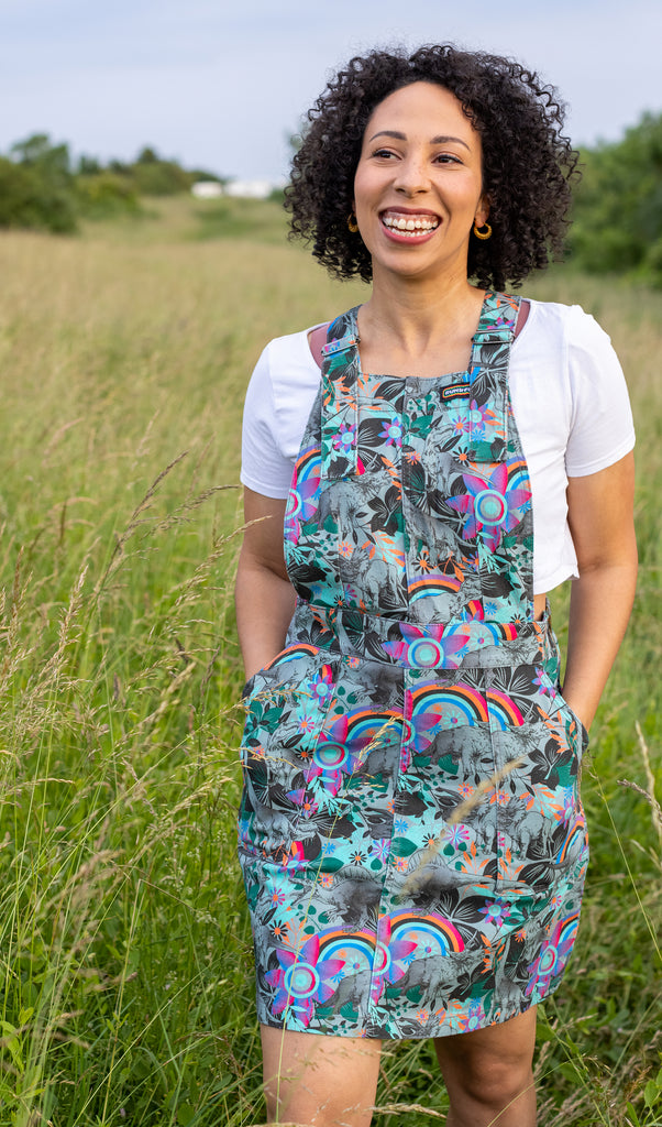 Amy is stood in a grass field wearing the jurassic dinosaur pinafore stretch twill dress with a crop white short sleeve tshirt underneath. They are facing the camera smiling looking off to the left with both hands in the front pockets. Photo is cropped from the knees up.