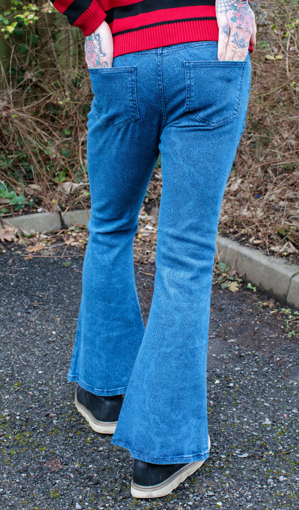 Jim is stood in a garden wearing the retro stretch stone wash denim Hendrix paisley bell bottom flares with a black and red striped jumper and black boat shoes. They are facing away from the camera with one leg forward to highlight the flares with both hands in the back pockets. Photo is cropped from the waist down.