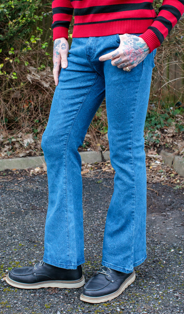 Jim is stood in a garden wearing the retro stretch stone wash denim Hendrix paisley bell bottom flares with a black and red striped jumper and black boat shoes. They are facing forward posing with one leg bent forward to highlight the flare shape.