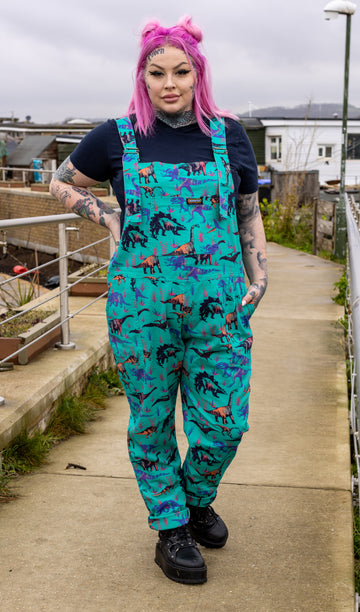 Zoe wearing the jade adventure dinosaur stretch twill dungarees with a short sleeve black tshirt and black boots. She is facing forward standing on a dock posing with one arm on her hip and the other in the dungaree pocket.