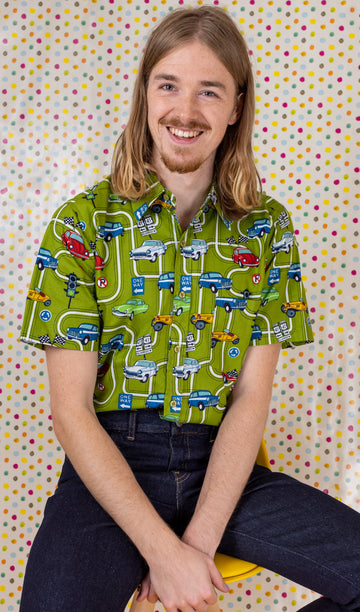 Jack is sat on a yellow stool in front of a multicoloured polka dot background wearing the kiwi car highway short sleeve shirt with blue denim jeans. They are looking into the camera and smiling with both hands in their lap. Photo is cropped from the thighs up.