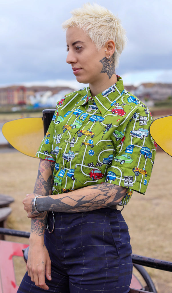 Yo is stood near a beach wearing the kiwi car highway short sleeve shirt with blue tartan trousers. They are facing the left with their hands crossed over in front of them. Photo is cropped from the hips up.
