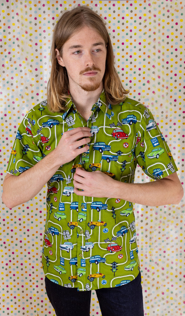 Jack is stood in front of a multicoloured polka dot background wearing the kiwi car highway short sleeve shirt with blue denim jeans. They are adjusting the buttons on the shirt and looking off to the right. Photo is cropped from the hips up.