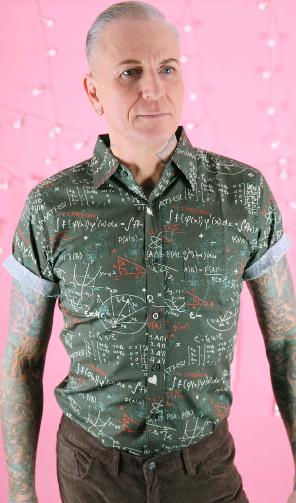Jim is stood in front of a pink studio background wearing the school chalkboard maths shirt with brown trousers. They are facing forward posing with both arms resting by their side whilst looking off to the right. Photo is cropped from the hips up.