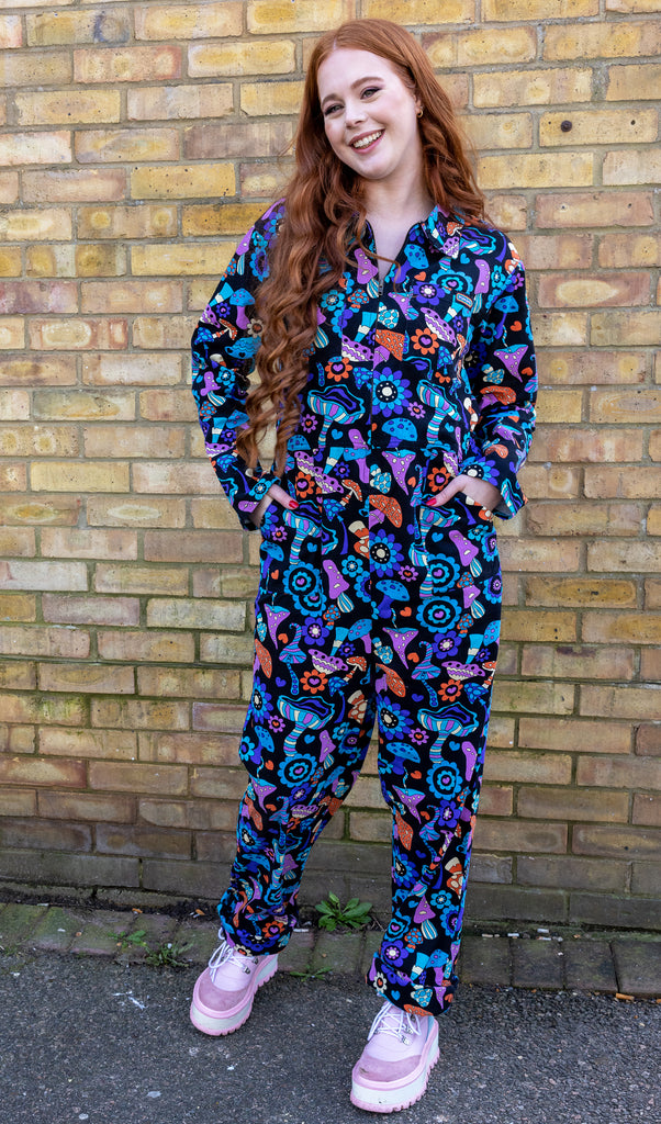 Sophie is standing by a brick wall wearing mushroom mayhem black blue purple orange pattern stretch twill boiler jumpsuit. Model is looking at the camera and smiling with hands in front pockets, jumpsuit has front zip and collared neck. Paired with pink platform boots