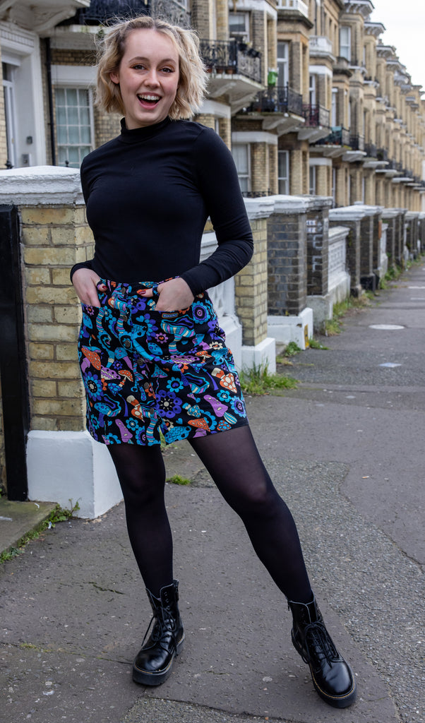 Amy is standing in front of a row of brick townhouses, wearing the mushroom mayhem a-line skirt with front button and high waist, paired with a black high neck long sleeve top, black tights and chunky boots. Model is looking at the camera and smiling with hands by their sides