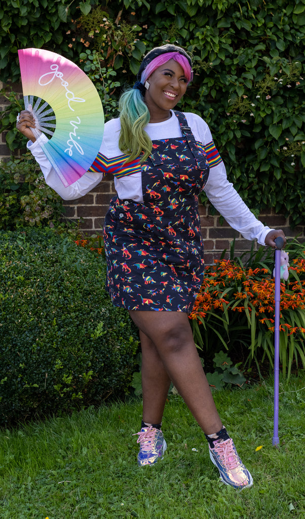 Ayesha is stood with her purple walking aid in a garden with one leg crossed over the other, she is looking off to the left and smiling. She is wearing the black rainbow dinosaur stretch twill pinafore dress with a white and rainbow stripe long sleeve t-shirt and holographic trainers. She is holding up a rainbow fan that says good vibes to match her rainbow hair.