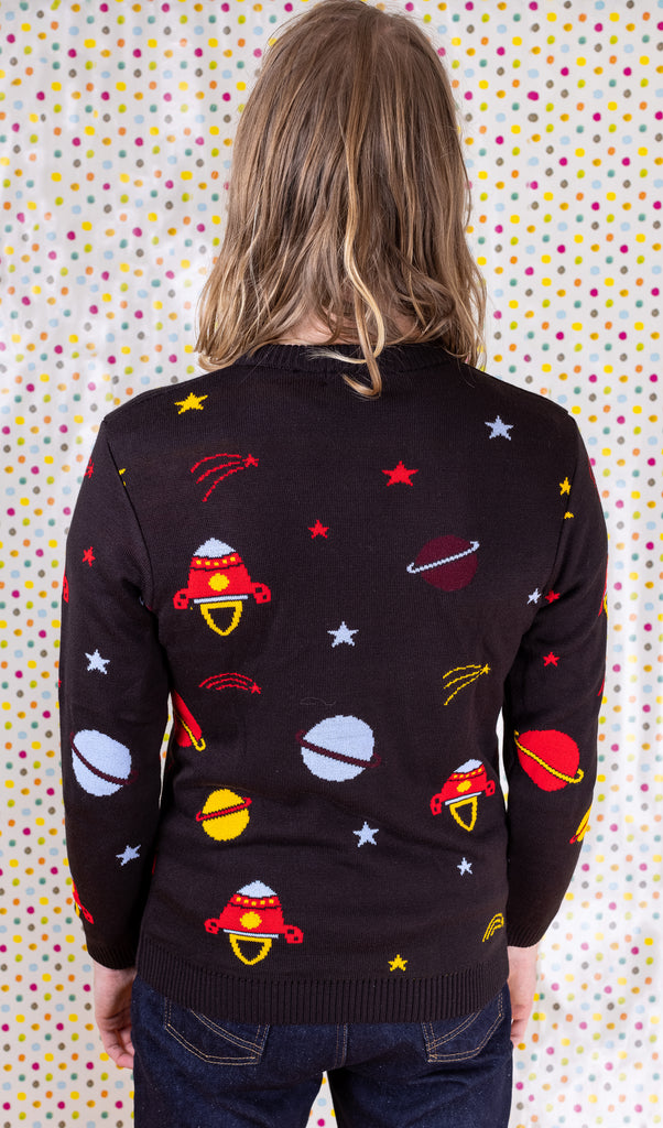 Jack is stood in front of a multicoloured polka dot background wearing the retro rocket outer space jumper with dark denim jeans. They are facing away from the camera to highlight the all over pattern on the jumper. Photo is cropped from the hips up.
