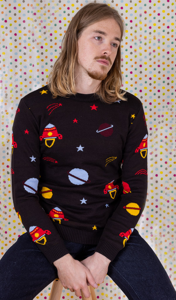 Jack is sat on a yellow stool in front of a multicoloured polka dot background wearing the retro rocket outer space jumper with dark denim jeans. They are facing forward with both hands clasped in their lap whilst looking off to the right. Photo is cropped from the hips up.