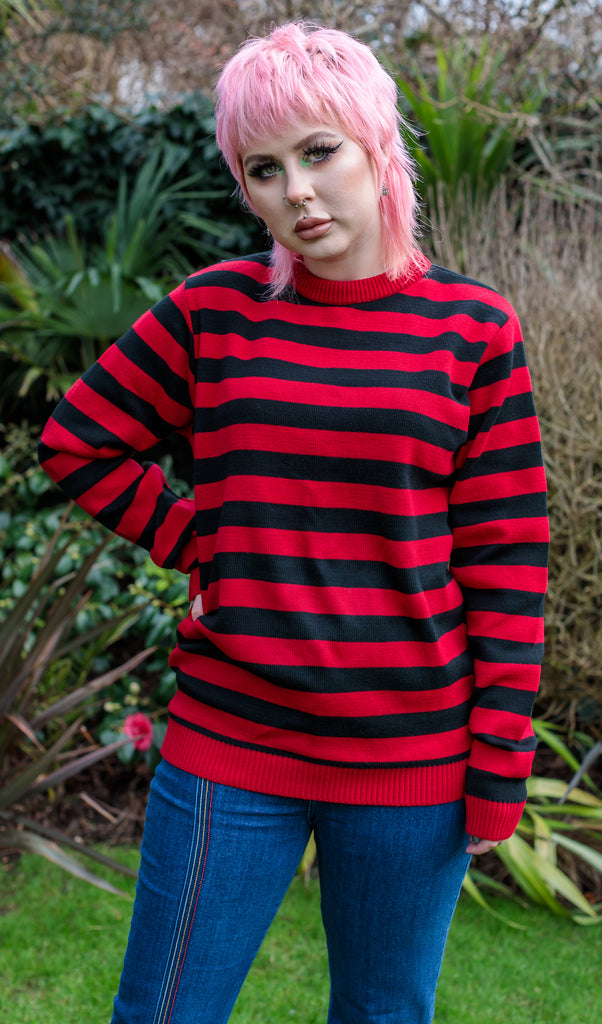 Florence is looking at the camera, standing outside. She has a pink mullet and is wearing the Red and Black Striped Jumper 