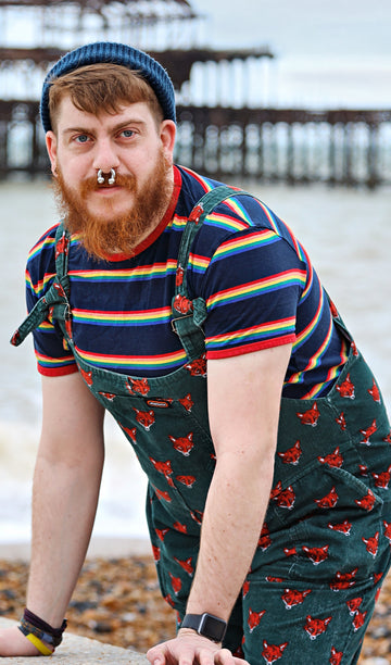 Model standing by the sea wearing a beanie, green fox dungarees and the Retro Rainbow Block Striped T Shirt.