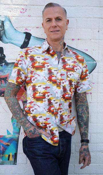Jim is stood in front of a graffiti wall wearing the retro sunset beach Hawaiian shirt with denim jeans. They're facing forward with one hand in their front jean pocket and smiling to camera. Photo is cropped from the knees up.