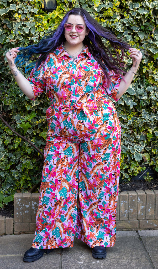 Luisa is wearing Pink Tiger Lily Stretch Jumpsuit with pink heart shaped glasses and black boots. The pink jumpsuit has an all over print of tigers and foliage. Luisa is stood in front of a leafy background facing the camera smiling with their hands in their blue and purple hair.