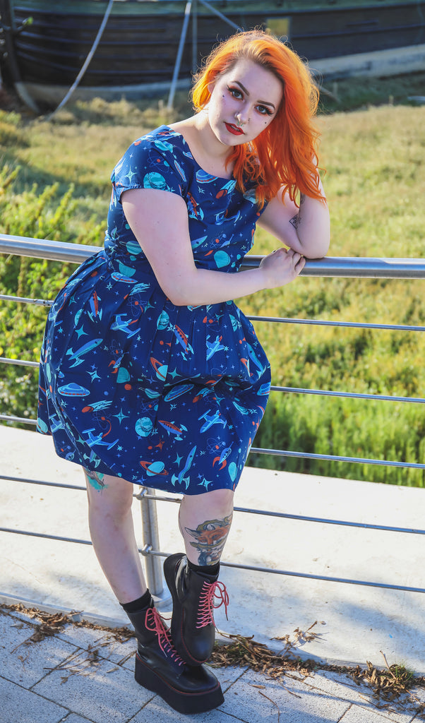 Tasha is stood in front of a river dock wearing the outer space retro tea party dress with black boots. She has mid length bright orange hair and tattoos. She is posing facing the camera leaning on the metal railing behind her resting her head on her hand. 